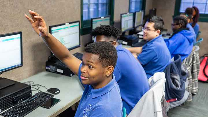 Secondary school student raising his hand while working at a computer.