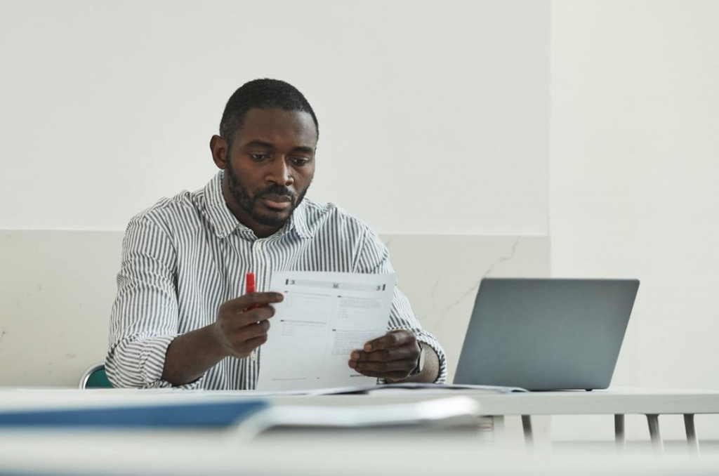 Black teacher reading a test paper at a desk.