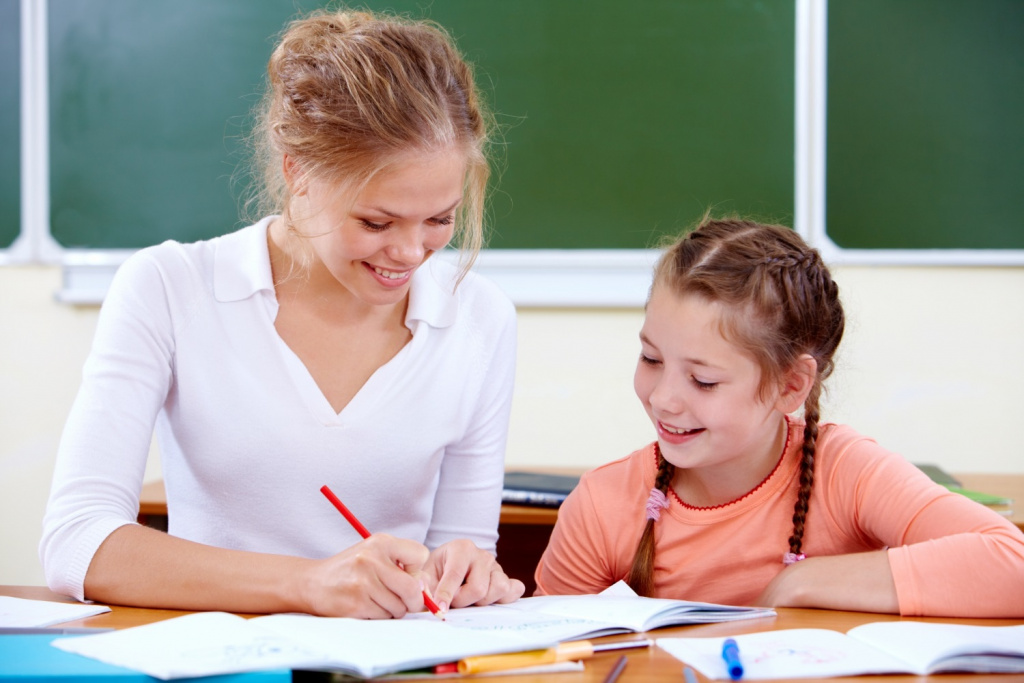 Elementary school teacher helping a girl write at her desk.