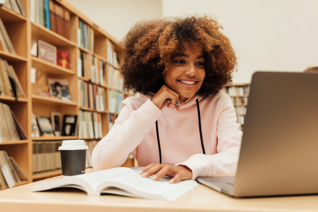 Black secondary school girl studying in a library with a laptop and textbook.