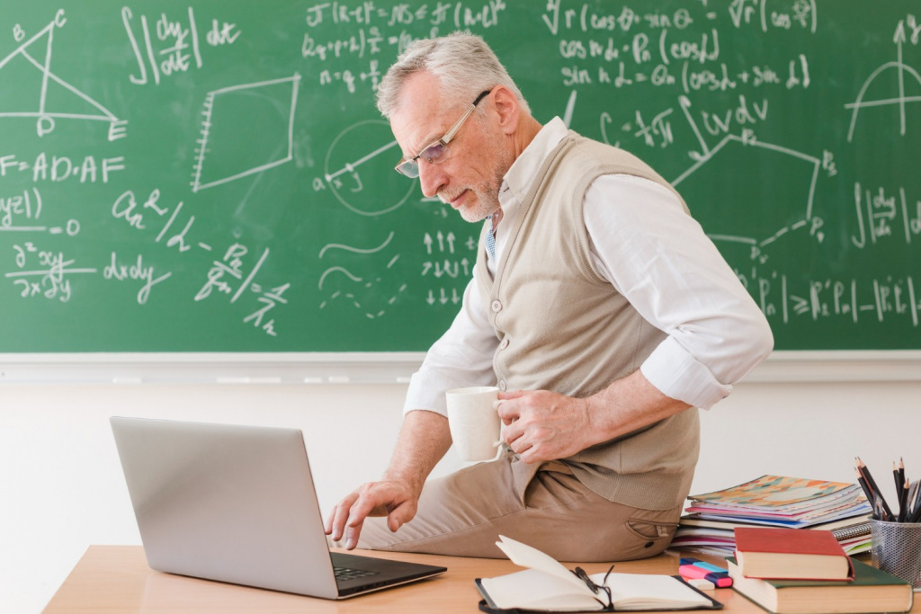 Older male teacher using a laptop in a classroom.